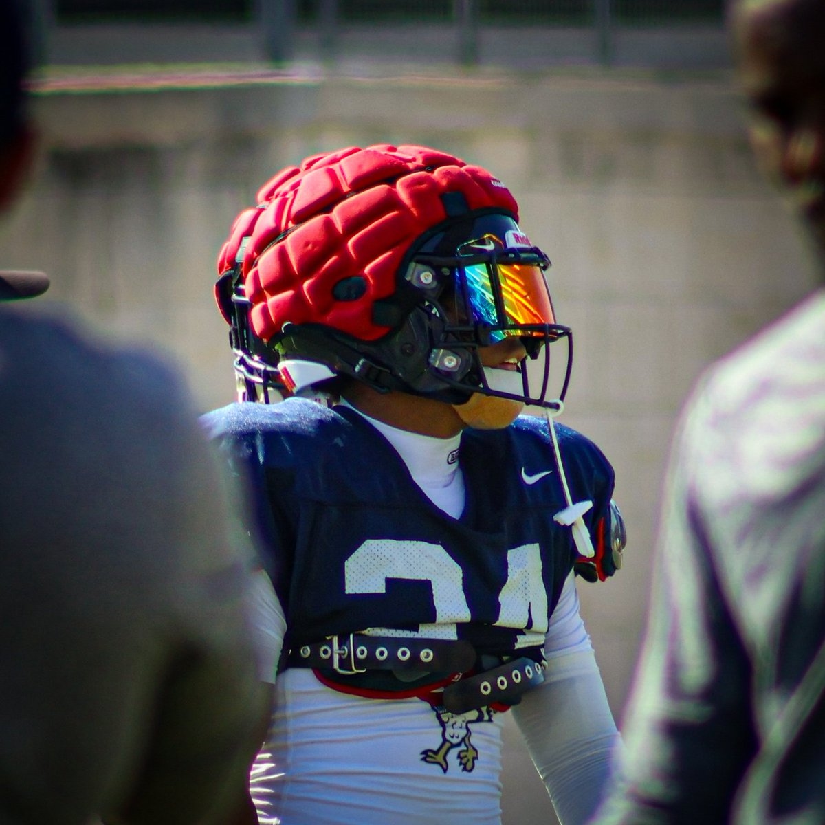 Young football player wearing red helmet and navy blue jersey with number 21, standing on sideline during game