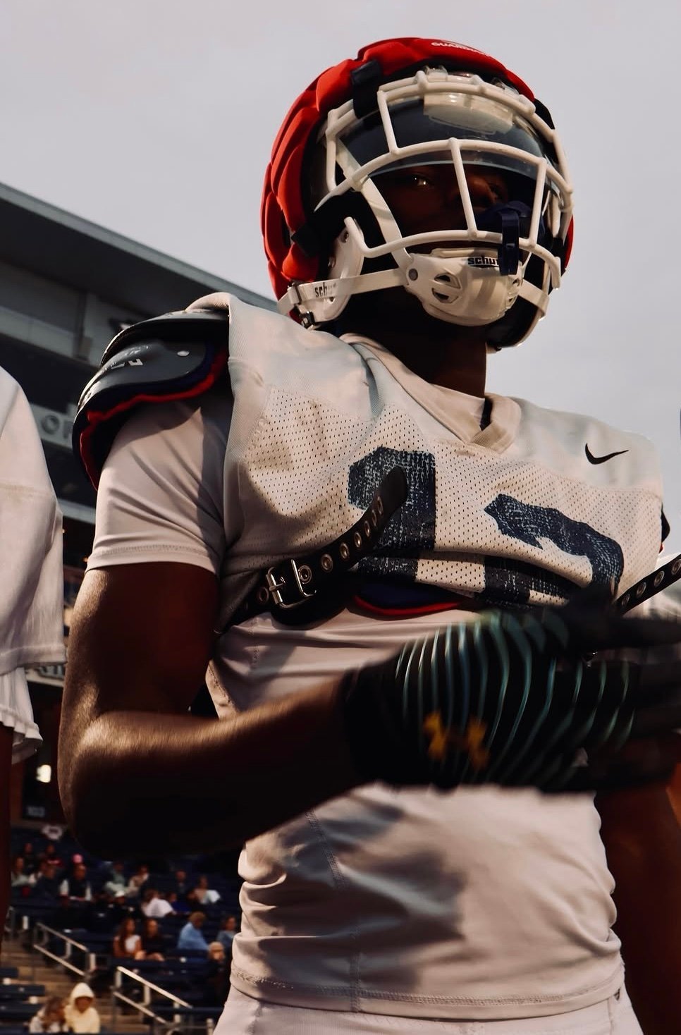 Football player wearing white jersey and red helmet with gloves, standing on sideline at stadium