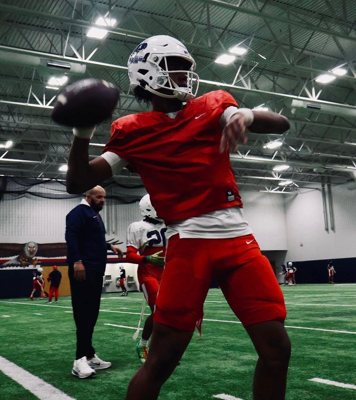 Football player in red uniform and white helmet throwing a pass indoors under bright stadium lights