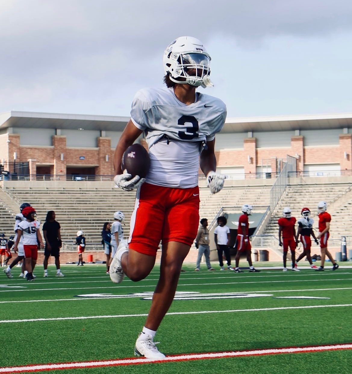 Young football player in white helmet and red shorts running with ball on stadium field during practice